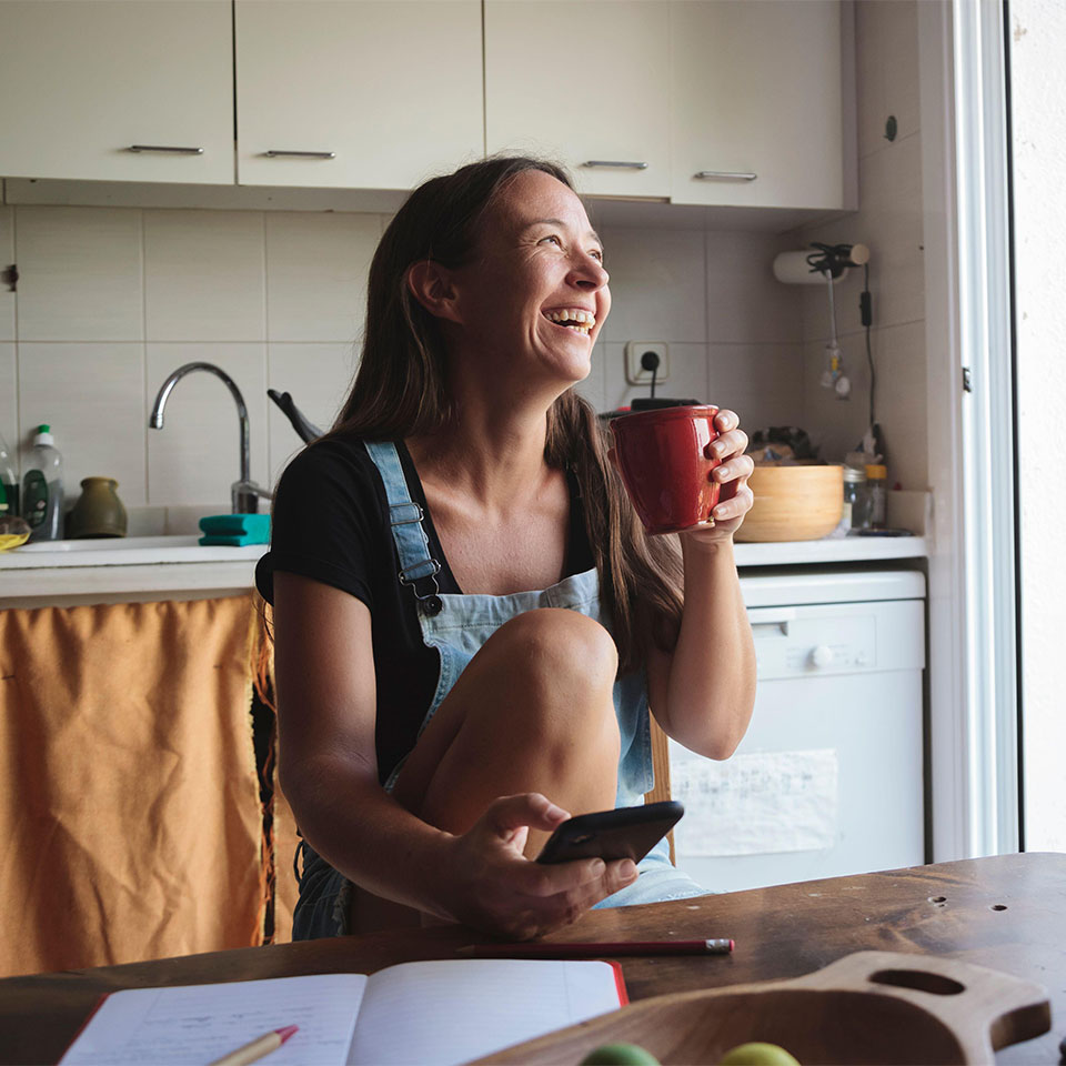 2C5449G - Young woman sitting on a wooden kitchen table with relaxed atmosphere checking her smartphone and writing notes; concept natural lifestyle, simplicity