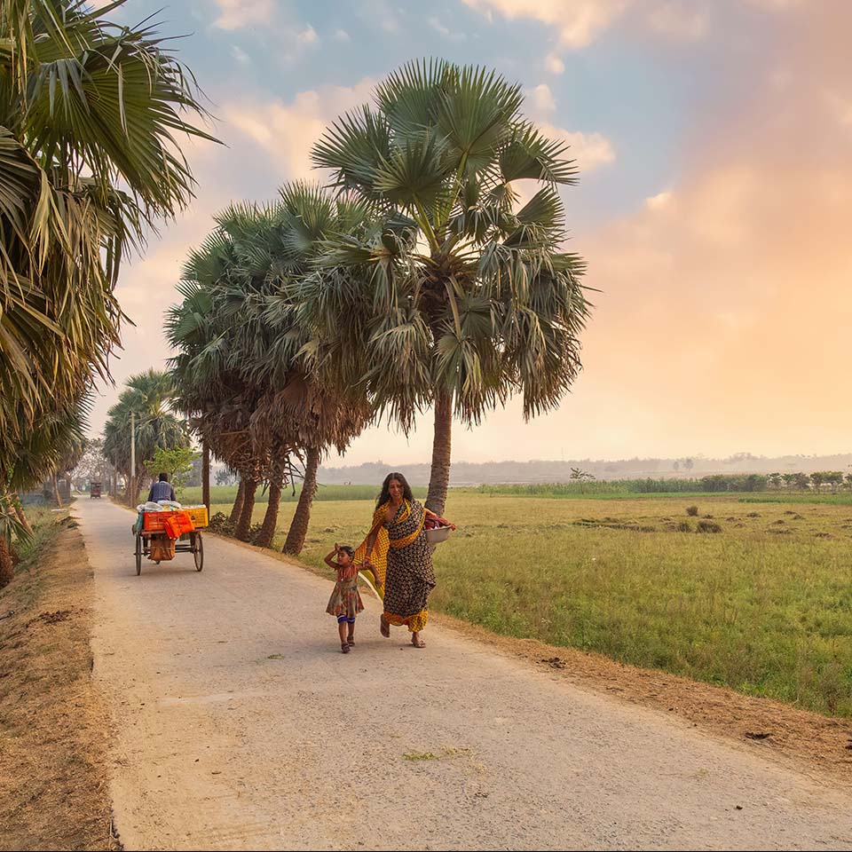 2EWTP9F - Rural woman with her kid walking down an unpaved village road with view of agriculture farmland at a district in West Bengal, India