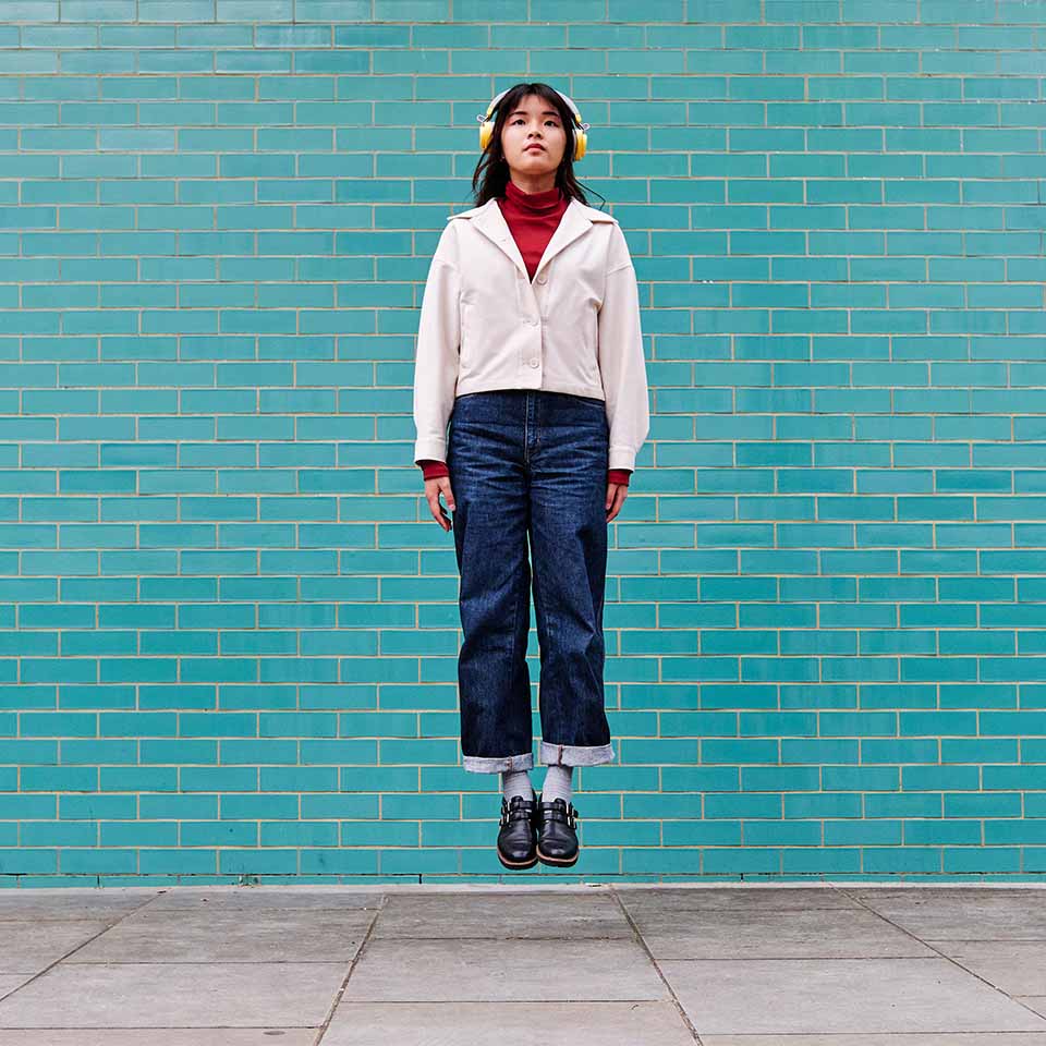 Woman with headphones levitating in front of turquoise brick wall